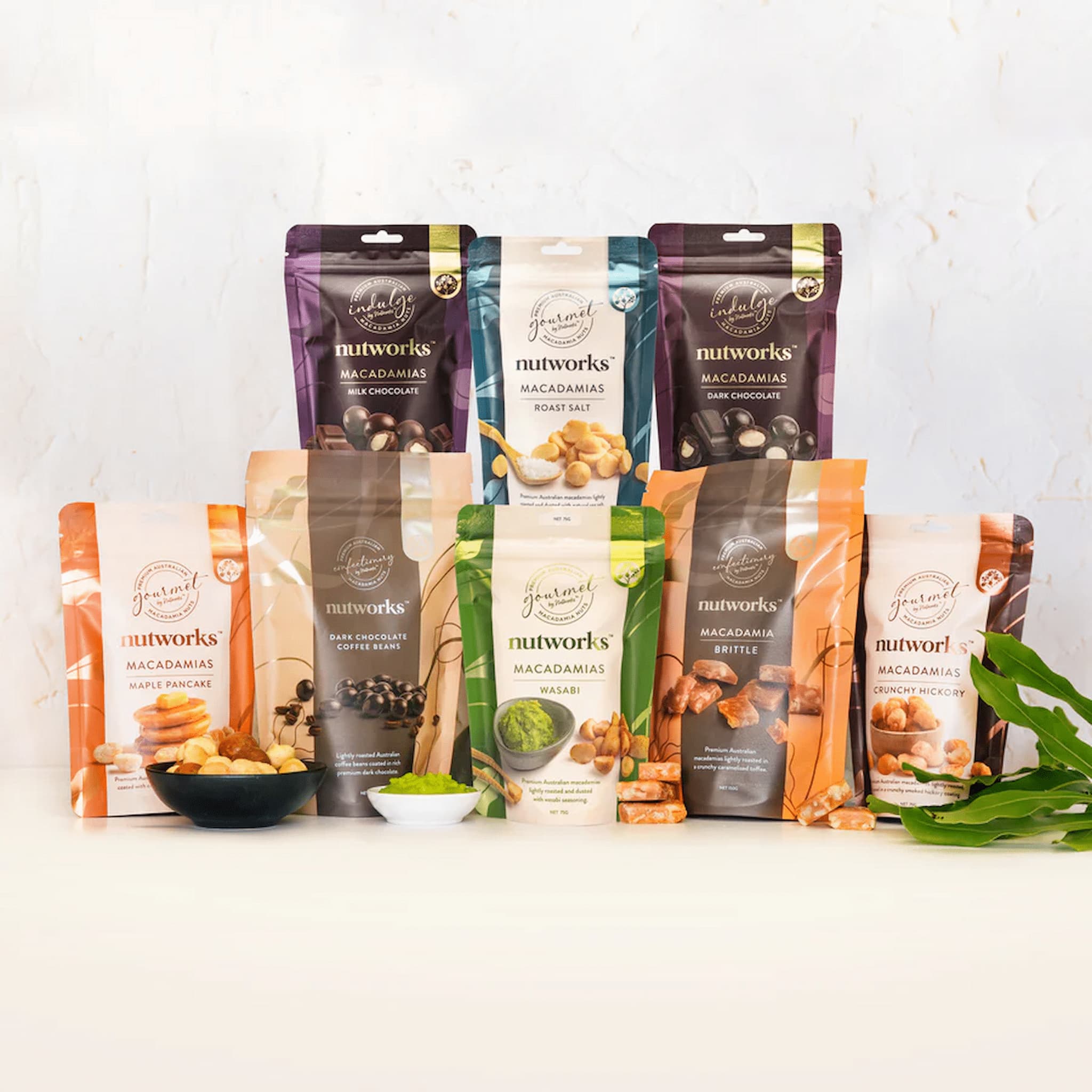 An assortment of packaged food items from Nutworks, including various macadamia nuts, savoury and sweet, in different flavours, arranged in a row on a white background.