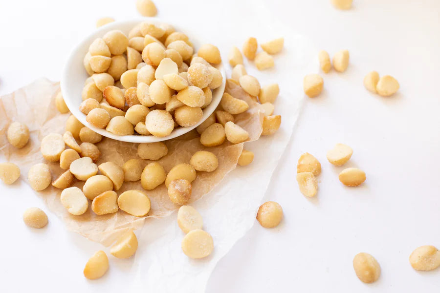 Roasted salted macadamia nuts scattered across a white surface alongside a small bowl of macadamias photo from Nutworks.